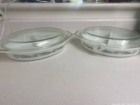 Pair of two white oval glass serving dishes with lids side-by-side on counter, one with black scroll decoration