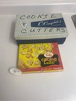 Photo of two boxed sets of cookie cutters. A blue-striped box labeled 'Cookie Cutters O'Comphies' behind a red and yellow box labeled 'Tala cookie cutters 12 attractive shapes'. Both boxes rest on a white surface.