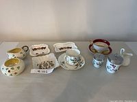 Overview photo showing all items in the lot including cups, saucers, cream pitcher, sugar bowl, decorative plates, lidded jar, and a small figurine. The items are displayed on a tabletop with clear visibility of shapes and styles.