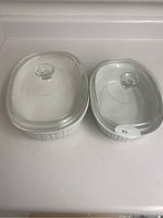 Two white ceramic rectangular serving dishes with clear glass lids placed side by side, showing ribbed exterior sides and round knob handles on lids.