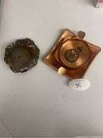 View of two coasters including a brown agate slice coaster and a copper-tone coaster with government crest decal and handles