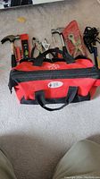 Hand tools and the red tool bag laid out on the carpet, showing a claw hammer, adjustable wrenches, channel lock pliers, utility knife, hex key set, spirit level, hand saw, pipe cutter, and a multi-tool pouch.