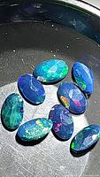 Close-up image of ten oval cut Ethiopian black opal gemstones with visible blue-green hues and rainbow flashes in a black dish.