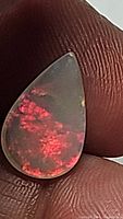 Front view of pear-shaped fire opal held between fingers showing vibrant red internal flashes and smooth polished surface.