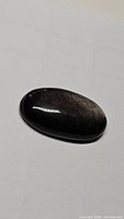 Polished oval cabochon silver sheen obsidian gemstone showing reflective silver flash on black background.