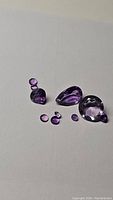 Photo showing eight natural purple amethyst gemstones of different cuts and sizes displayed on a white background.