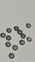 Photo showing thirteen round mystic topaz gemstones with a greenish reflective color on a white background.