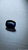 Single cushion-shaped natural blue sapphire with green undertone, shown on light background under varied lighting.