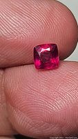 Close-up of ruby gemstone held between fingers, showing deep vivid red pink color and faceted cut.