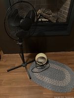 Photo showing two fans (a black oscillating pedestal fan and a small cream tabletop fan) next to an oval braided rug on floor.