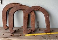 Three rusted metal horseshoe hammers on a wooden shelf with a measuring tape showing their size.