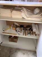 Image showing a white shelving cabinet with folded white drop cloths on top shelf and various paintbrushes and roller covers on bottom shelf.