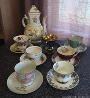 Photo of SB&Son coffee pot and seven assorted teacups with saucers on a table showing the shape and floral details of the coffee pot and variety of cup designs.