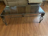 Front angled view of the wrought iron and glass coffee table on a wooden floor showing the decorative iron base and rectangular glass top.
