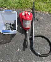 Overall view of the red Dirt Devil Featherlite Cyclonic vacuum with hose and wand attachments beside a clear plastic container holding additional vacuum attachments, placed on asphalt with grass background.