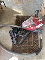 Red Dirt Devil canister vacuum on a brown doormat with hose and multiple cleaning attachments visible, vacuum body dusty and used.