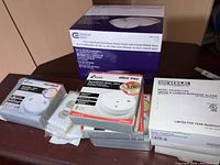 Stack of various smoke and carbon monoxide alarms from Kiddie, Knauf, and Universal brands alongside white outlet covers
