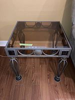 Square wrought iron framed end table with tinted glass top positioned on wood flooring against a wall.