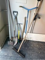Photo showing six gardening tools arranged upright against a wall on a floor: edger, spade, rake, axe, hoe/pick, claw twister.