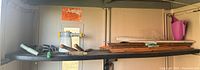 Wide angle photo showing a variety of gardening tools and supplies on a metal shelf inside a shed. Items include a sprayer, hand tools with wooden handles, pruning shears, small fencing segments, and a pink watering jug.