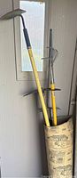 Photo of three gardening tools (two cultivators and one hoe) standing inside a container against a white wall with a window behind them.