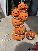 Photo of six orange plastic jack-o'-lanterns stacked vertically with expressive faces, connected by power cord, next to one small pumpkin figurine with a painted face.