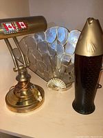 Photo showing three items: a dark olive fish-shaped glass bottle with gold-tone metal top, a vintage brass desk lamp with Canadian flag stickers, and an art deco fan-shaped table lamp.