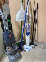 Full view of all cleaning items against basement wall including Hoover Turbo Scrub, brooms, mops, and handheld steamer