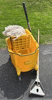 Full view of the yellow Rubbermaid mop bucket with mop and handle resting inside it, showing wheel caster and mop head.
