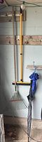 Full view of the lawn rake, rubber floor scrubber, and two umbrellas hanging on a wall.
