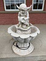Full view of ceramic garden fountain with cherub figure holding a shell, scalloped water basin, shown outdoors on a tiled surface.