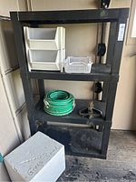 Black plastic shelving unit holding white plastic bins, green water hose, metal sprinkler, and styrofoam box labeled grass seed