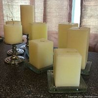 Photo showing collection of five square pillar candles of varying heights resting on clear glass square plate holders on a dark surface with a window curtain background.