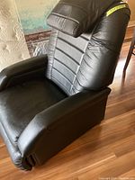Side view of black leather-like recliner chair showing padded armrest, headrest pillow, and power cable on side.