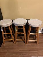 Front view of three wooden stools with light-colored upholstered round seats arranged side by side on wood floor against beige wall.