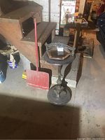 Photo showing the floor stand ashtray beside the red metal snow shovel against a stair background.