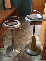 Photo showing two vintage ashtray stands with metal bases and amber glass ashtrays installed, placed on a carpeted floor near wood paneling and furniture.