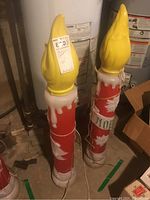 Two electric Christmas candle decorations with yellow flame tops and red and white bodies placed on concrete floor near pipes and cardboard boxes