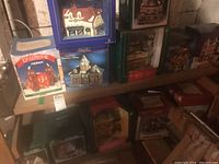 Photo showing multiple boxed Christmas village houses and shops on shelves, including a Noma porcelain lighted house under the Dickensville Collectables brand.