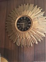Front view of gold tone sunburst wall clock on wood panel wall.