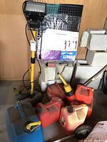 View of the work lamp on rolling stand alongside six assorted empty gas cans of various colors and sizes.