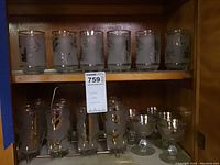 Full shelf showing six tall frosted glasses with gold leaf appliqué on the top shelf, six dessert cups with stems on the bottom shelf along with a glass holder.