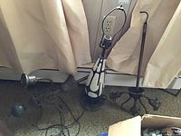 Photo of a Mid-Century Modern style table lamp with an abstract black and white design alongside two metal chandelier fixtures with multiple sockets, placed on carpet in front of beige curtains.