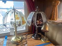 Two vintage decorative desk lamps on a wooden surface by a window, one with black base and carousel horse panels, the other with brass base and angel panels, some panels missing on angel lamp.