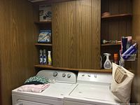 Laundry room setup with washer, dryer, wooden cabinets, laundry cleaners and household items stored on shelves above and around machines.