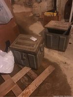 Two metal military boxes placed on a wooden surface in a basement-like setting, showing their top and side views.