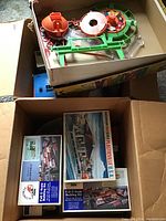 Box containing loose plastic parts of a Hoopla game and stacked Plasticville USA building kits boxes.