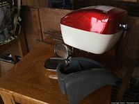 Red and white hard saddlebag on a wooden surface with motorcycle mirrors and black fender below it.