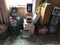 Multiple vintage musical carousel boxes stacked near window with curtain, some decorative boxes and a framed picture on floor.