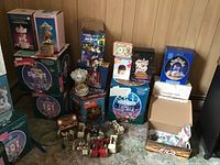 Overview of multiple boxed musical carousels, decorative clocks, string light components, and a porcelain doll, arranged on floor against wood-paneled wall.